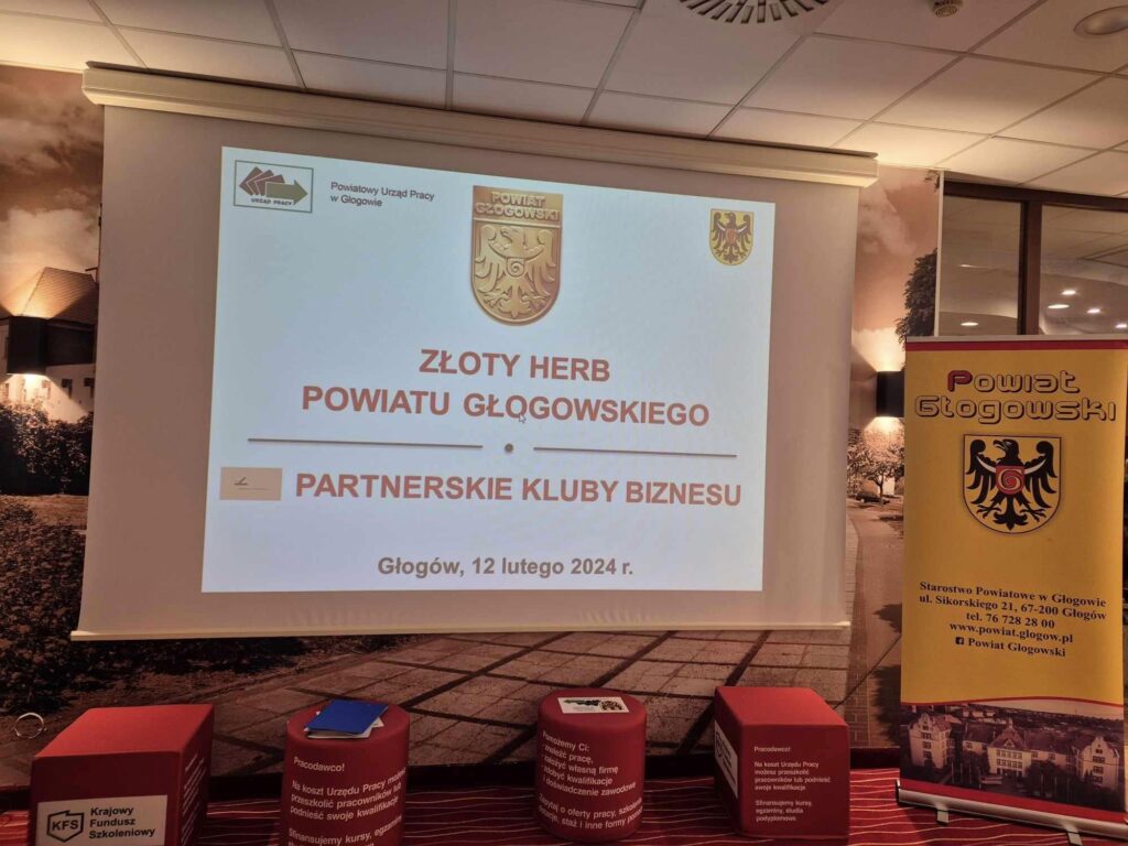 Zdjęcie przedstawia prezentację na konferencji rozdania złotych herbów. Treść przedstawiona na slajdzie: Złoty Herb Powiatu Głogowskiego Partnerskie Kluby Biznesu. Głogów, 12 lutego 2024 rok