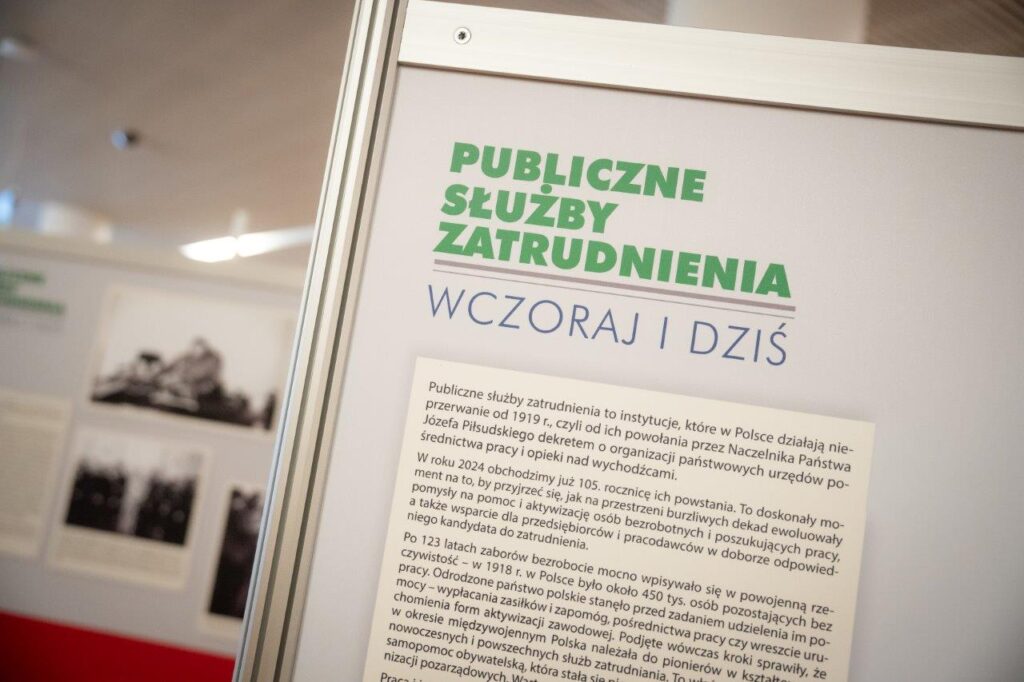 Zdjęcie zawierające cząstkową informację o treści: Publiczne Służby Zatrudnienia wczoraj i dziś: Publiczne służby zatrudnienia to instytucje, które w Polsce działają nieprzerwanie od 1919 roku od ich powołania przez Naczelnika Państwa Józefa Piłsudskiego dekretem o organizacji państwowych urzędów pośrednictwa pracy i opieki nad wychodźcami. W roku 2024 obchodzimy już 105 rocznicę ich powstania. To doskonały moment na to, by przyjrzeć się jak na przestrzeni burzliwych dekad ewoluowały pomysły na pomoc i aktywizację osób bezrobotnych i poszukujących pracy, a także wsparcie dla przedsiębiorców i pracodawców w doborze odpowiedniego kandydata do zatrudnienia. Po 123 latach zaborów bezrobocie mocno wpisywało się w powojenną rzeczywistość - w 1918 roku w Polsce było około 450 tys. osób pozostających bez pracy. Odrodzone państwo polskie stanęło przed zadaniem udzielenia im pomocy - wypłacania zasiłków i zapomóg, pośrednictwa pracy czy wreszcie uruchomienia form aktywizacji zawodowej. Podjęte wówczas kroki sprawił, że w okresie międzywojennym Polska należała do pionierów (...)