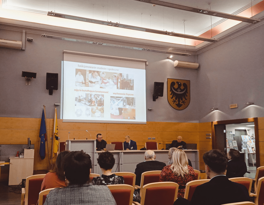 Zdjęcie przedstawia uczestników konferencji siedzących tyłem wraz przedstawicielami Polskiego Towarzystwa Walki z Kalectwem, którzy znajdują się na scenie.